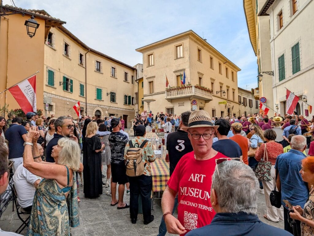 A crowd filling the medieval square at Chianciano Terme for the Biennale opening, flags strung across the buildings