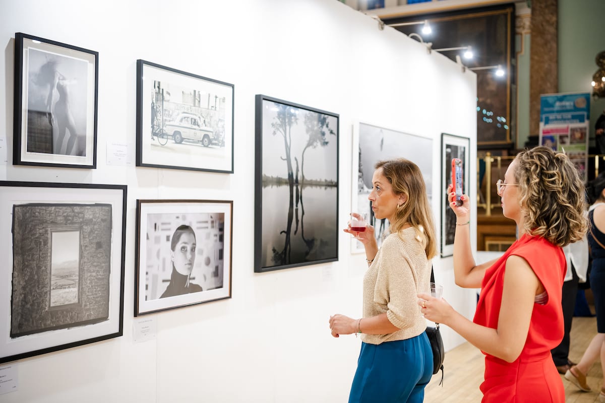 Two visitors with drinks looking along a wall of framed photographs at the London Art Biennale