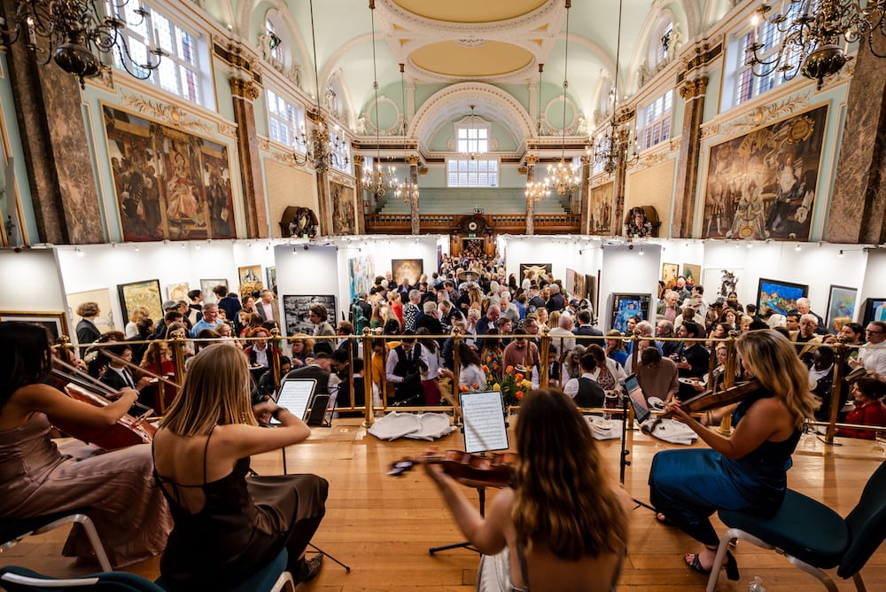 A string quartet playing to a full room at the London Art Biennale, Chelsea Old Town Hall, paintings hung the length of the hall behind them